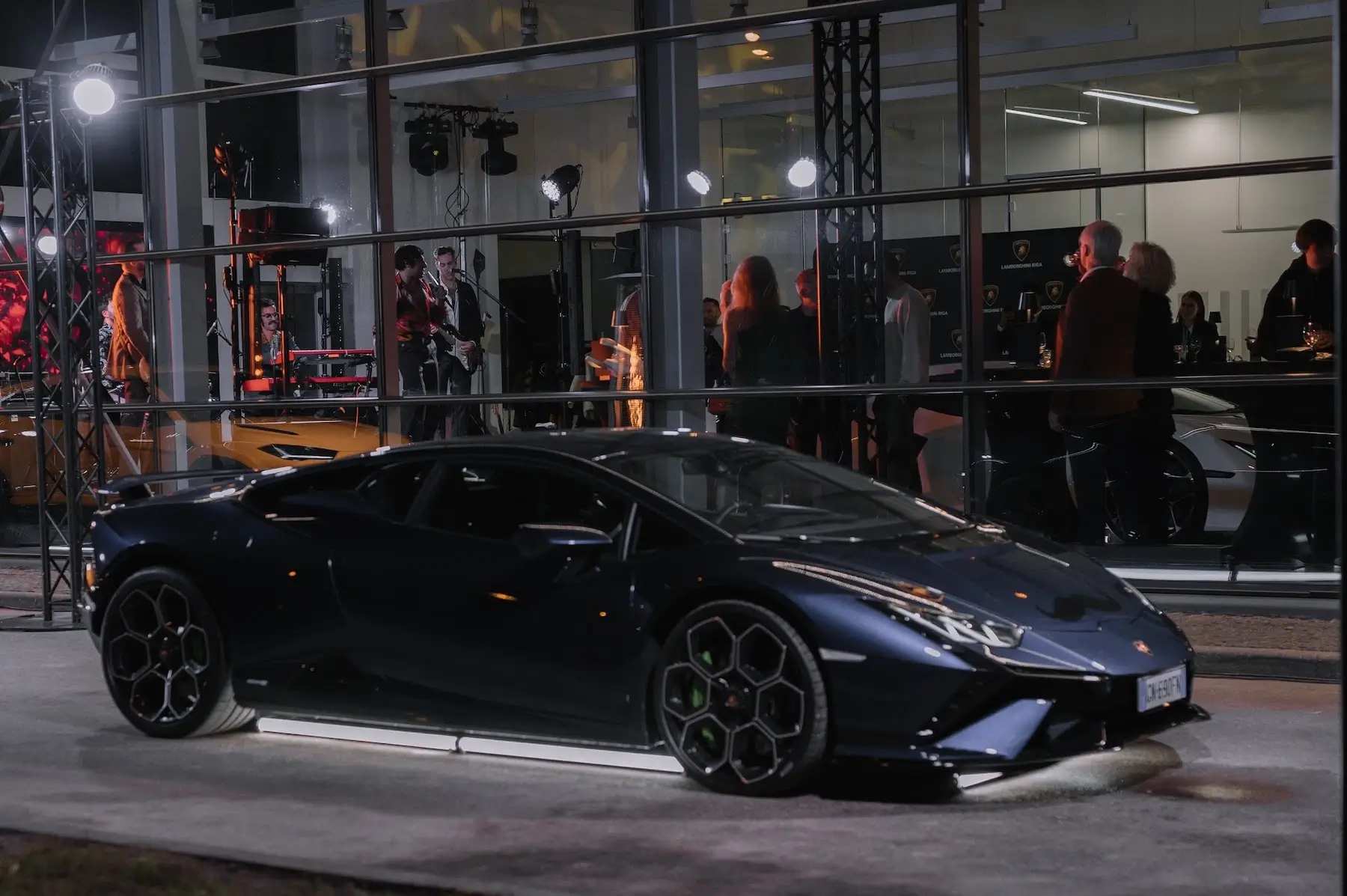 Lamborghini sporta automašīna izstādīta vakara pasākumā stiklotā telpā ar apmeklētājiem fonā