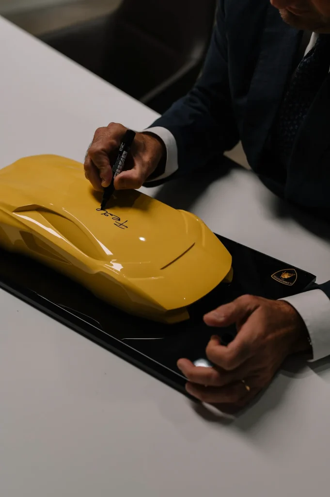 Vīrietis parakstās uz dzeltenas Lamborghini automašīnas formas dekorācijas uz galda