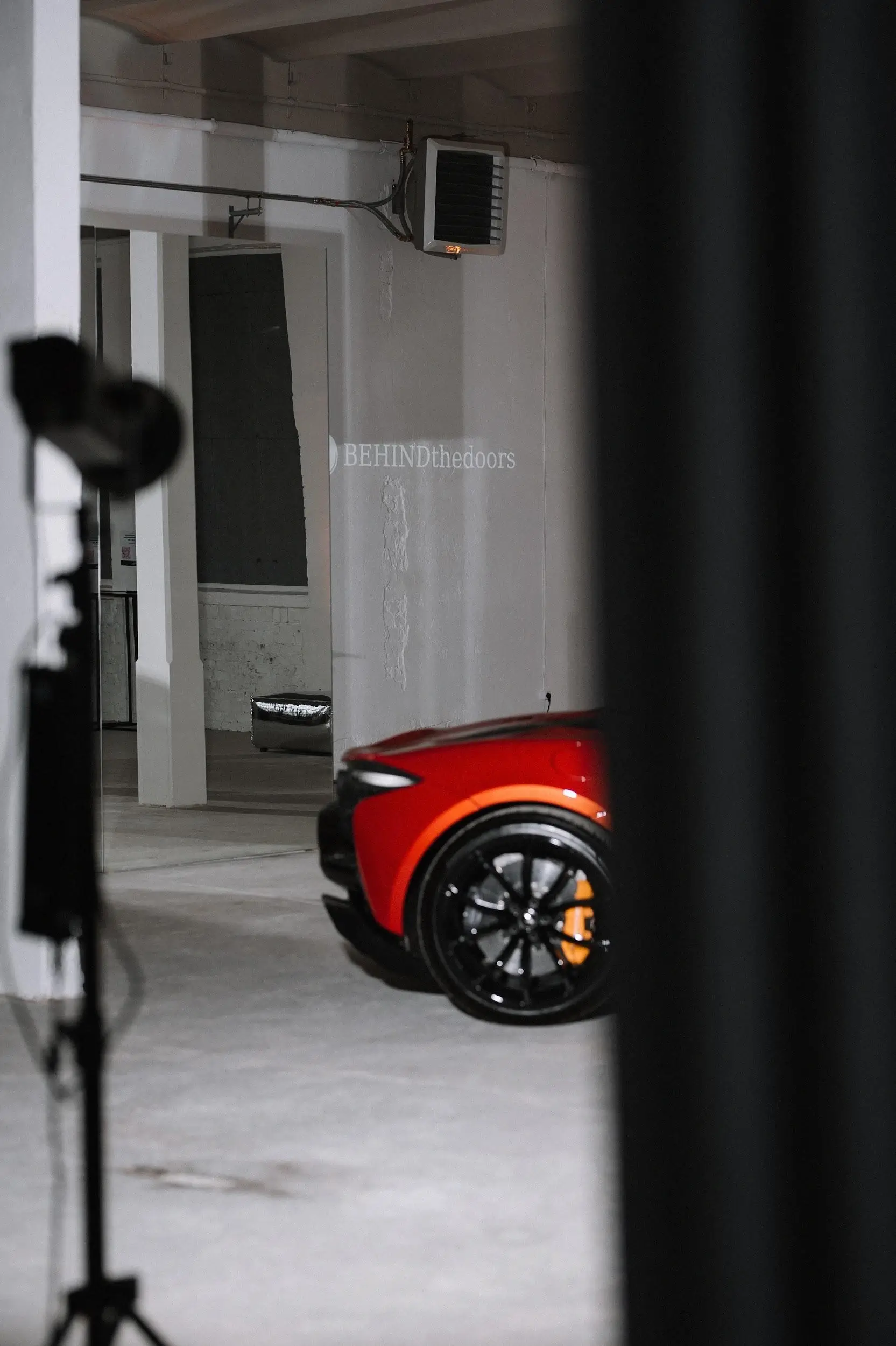 A red sports car in a spacious hall during a photoshoot, with the "BEHINDthedoors" logo visible on the wall.