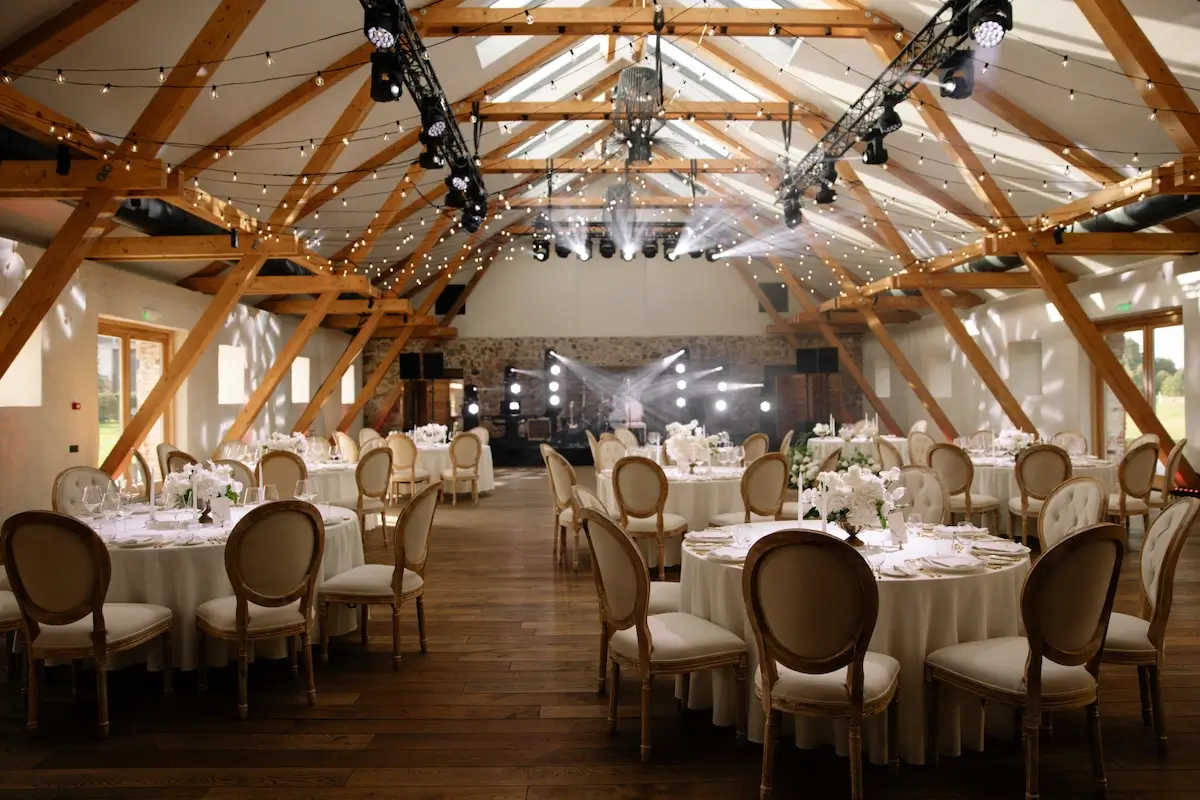 Elegantly set tables in an event hall with wooden beams, lighting, and a stage in the background