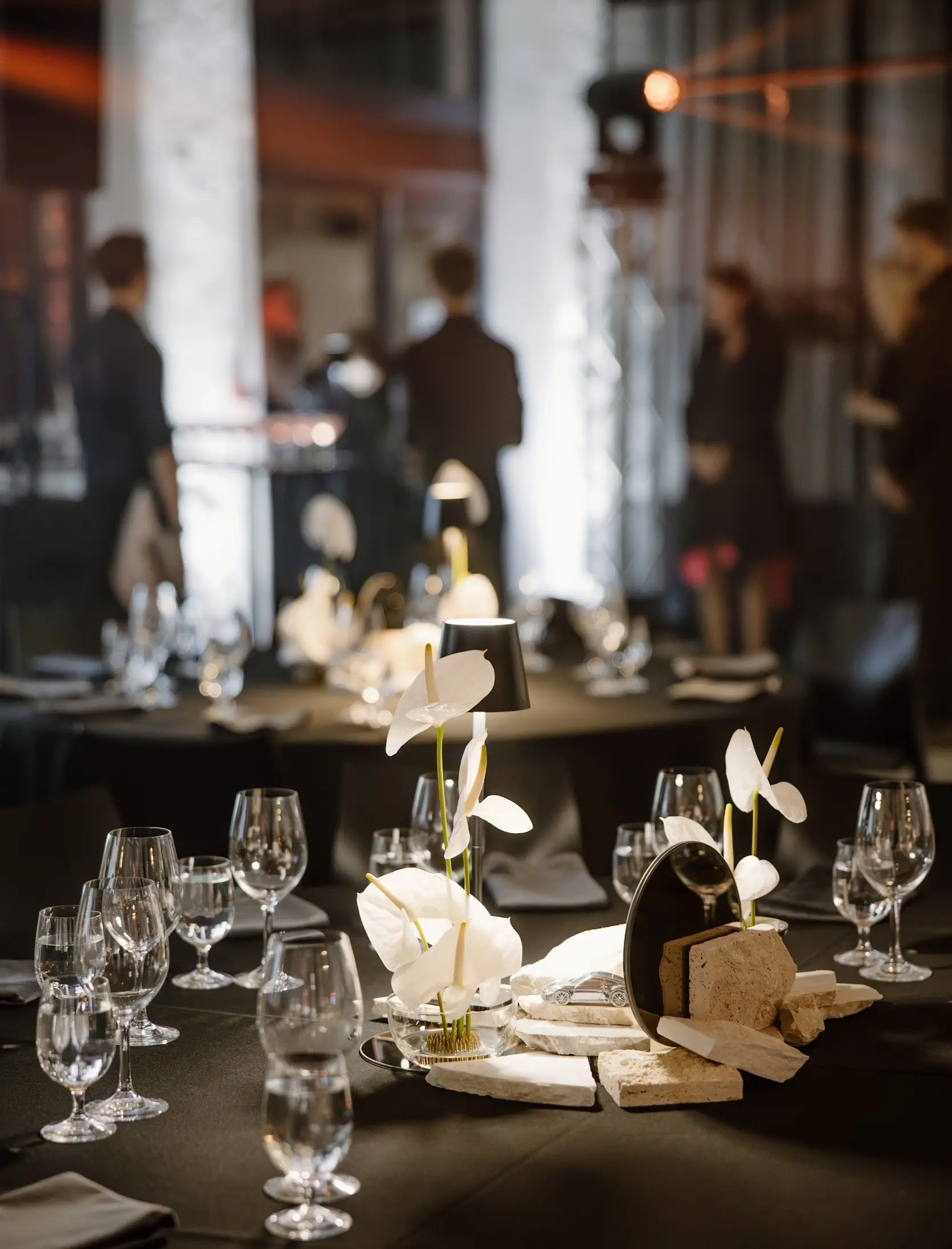 Elegantly set tables with glasses, white flowers, and decorative stone elements in a spacious hall with guests in the background.