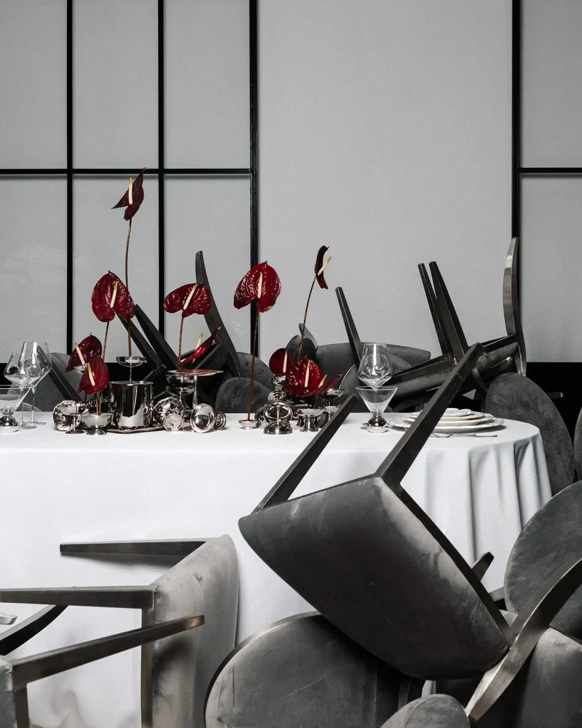 An elegant table setting with a white tablecloth, silver dishes, and red flower decorations in a spacious hall.