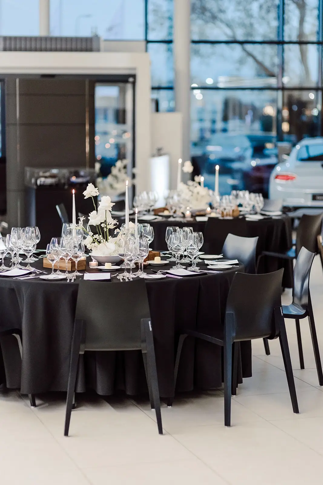Round tables with black tablecloths, glasses, candles, and flower arrangements in a room with large windows