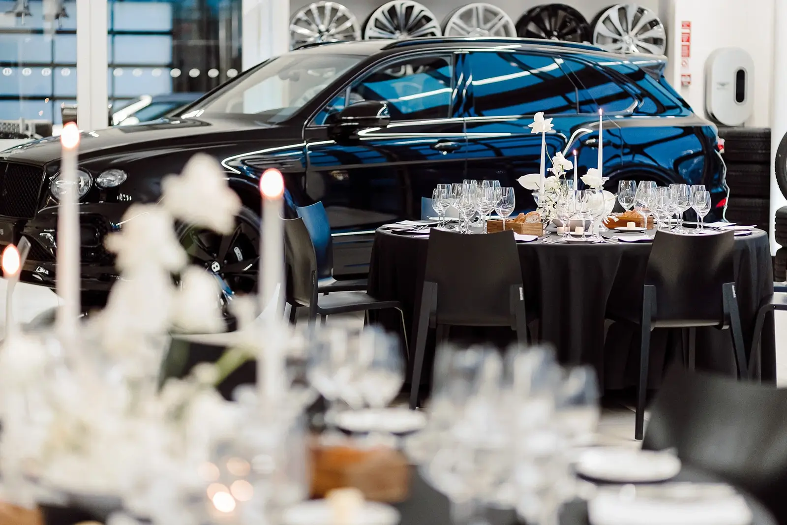 Table setting with black tablecloths, glasses, candles, and flower arrangements in a room with a car in the background
