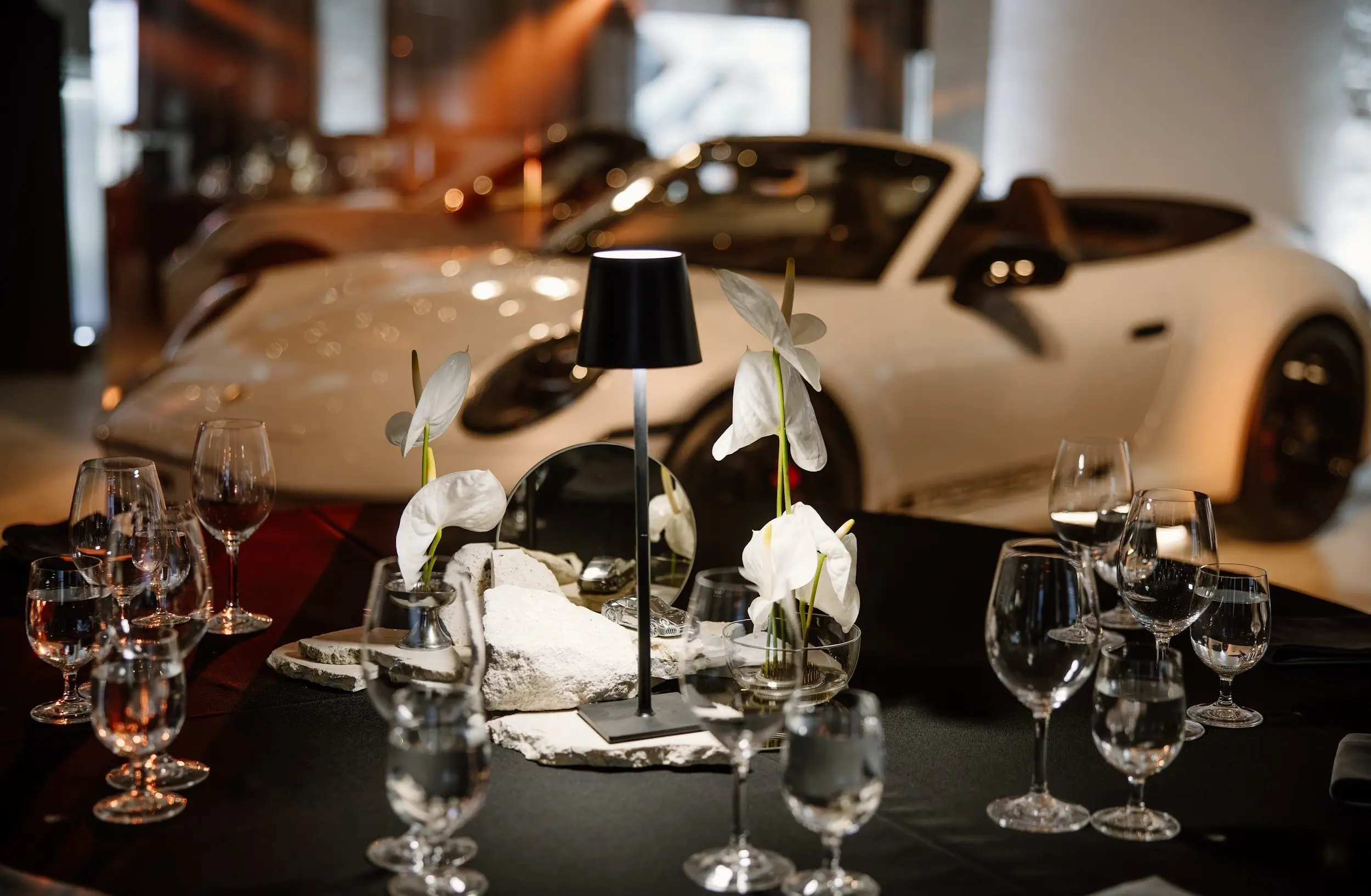 An elegant table setting with glasses, white flowers, and a table lamp, with a sports car in the background in a spacious hall.