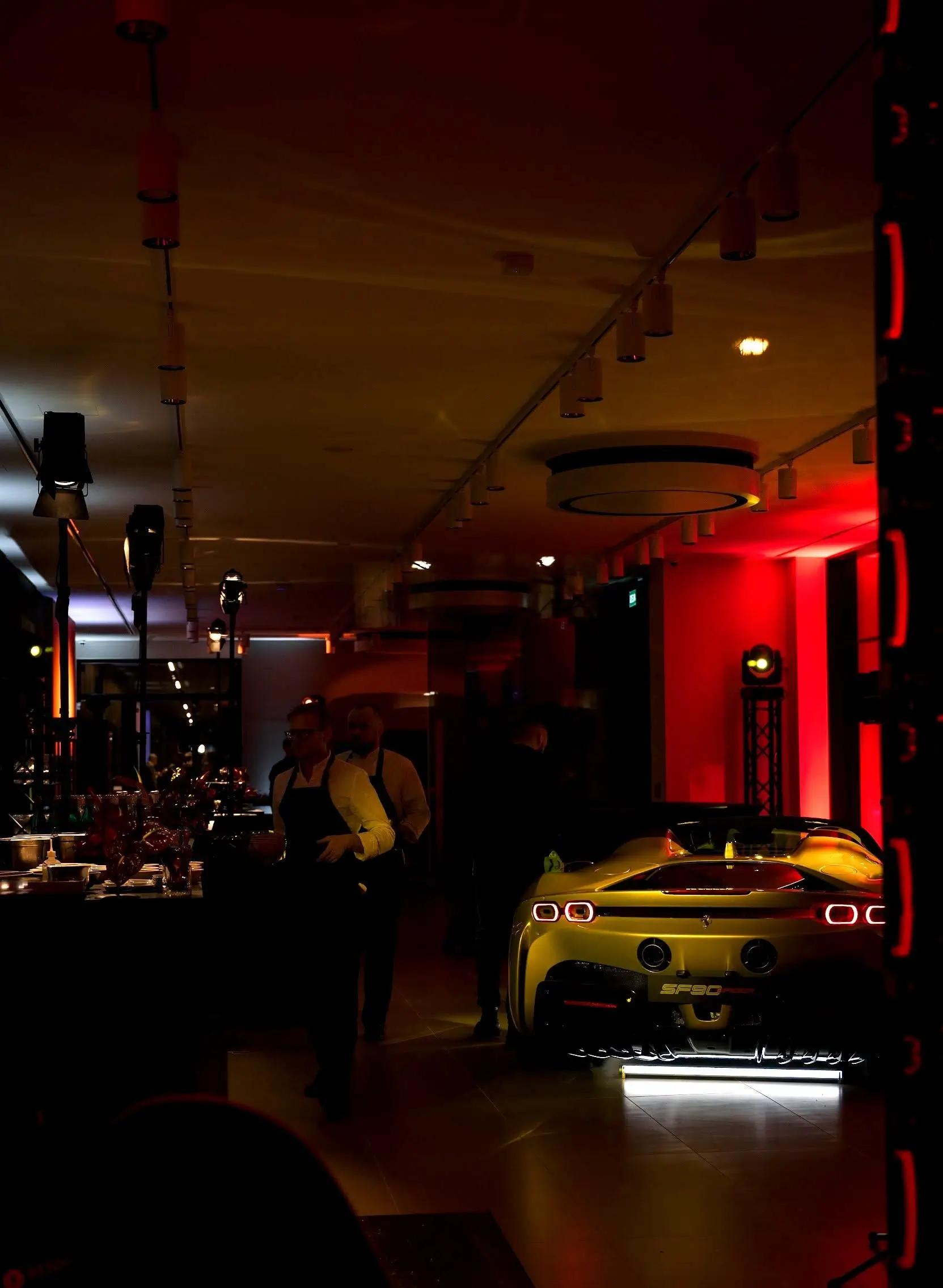 A yellow sports car in a room with red lighting, waiters, and a counter in the background